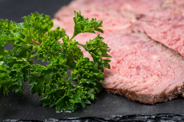 Sliced Grass Fed Juicy Corn Roast Beef garnished with Fresh Curly Parsley on black natural stone background.