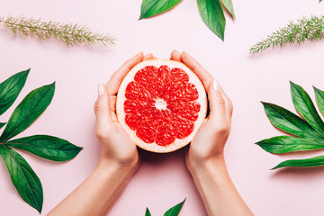 Female's hands holding grapefruit on pink background with tropic plants and leaves.