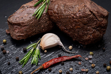 Grass Fed Corn Roast Beef garnished with Fresh Rosemary, dried Red Chile Pepper, Garlic and Rainbow  Peppercorns on natural black stone background.