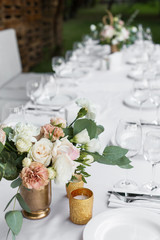 Wedding table setting decorated with fresh flowers in a brass vase. Wedding floristry. Banquet table for guests outdoors with a view of green nature. Bouquet with roses, eustoma and eucalyptus leaves