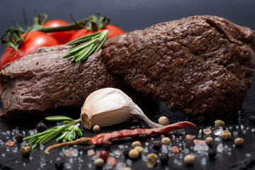 Grass Fed Corn Roast Beef garnished with Tomatoes, Fresh Rosemary, dried Red Chile Pepper, Garlic and Peppercorns on natural black stone background.