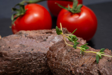 Grass Fed Juicy Corn Roast Beef garnished with Red Tomatoes and Fresh Thyme Herb  on black natural stone background.