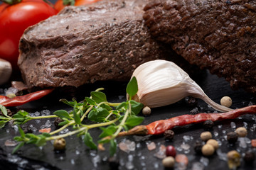 Grass Fed Corn Roast Beef garnished with Tomatoes, Fresh Thyme, dried Red Chile Pepper, Garlic and Peppercorns on natural black stone background.