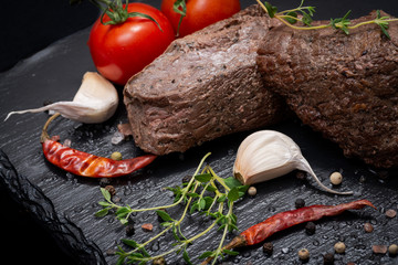 Grass Fed Corn Roast Beef garnished with Tomatoes, Fresh Thyme, dried Red Chile Pepper, Garlic and Peppercorns on natural black stone background.