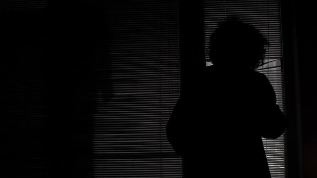 Sleepless lonely woman silhouette looking at outdoors lights over window blinds at night