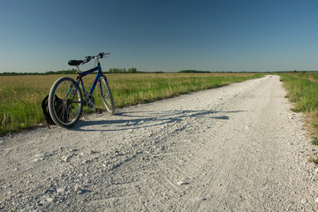 Obraz premium Standing bicycle and backpack on a gravel road to the horizon, green meadow and blue sky. Srebrzyszcze, Poland