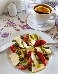 Soft cheese sliced into a large with spicy green seasoning and a little bit olive oil. Ripe red tomatoes. Cup tea with lemon in background. Meal served on a white utensils