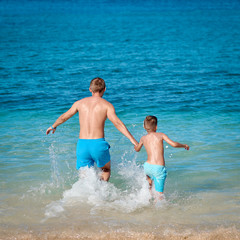 European boy and his dad are running in to the ocean holding hands, they are making a lot of splashes around.
