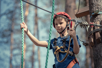 Child in forest adventure park. Kid in red helmet and blue t shirt climbs on high rope trail. Agility skills and climbing outdoor amusement center for children. Young boy plays outdoors. - 274124121