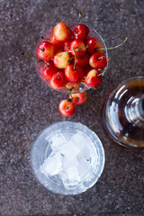 Yellow sweet cherries in a whiskey glass, ice cubes and a bottle of whiskey against a stone background. A refreshing cold cocktail at the base of the whiskey.