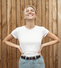 Young pretty girl is standing on a light wooden wall background. Woman is wearing a white empty...