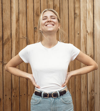 Young pretty girl is standing on a light wooden wall background. Woman is wearing a white empty t-shirt without logo which makes it being perfectly suitable for mock-up.