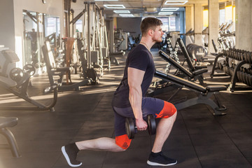 Side view portrait of concentration young adult sport man athlete working out in gym, standing on the knees and holding two dumbbells, doing exercises for legs and squatting. indoor, looking at camera