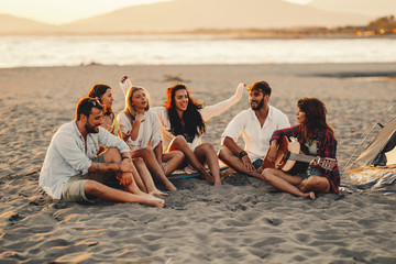 Happy friends sitting on the beach singing and playing guitar during the sunset