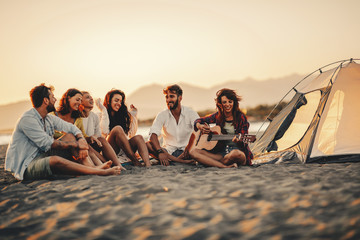 Happy friends sitting on the beach singing and playing guitar during the sunset - Powered by Adobe