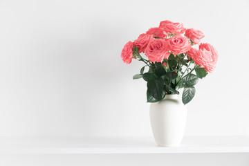 Roses flowers of coral color in vase on shelf against white wall. Space for text