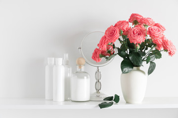 Soft light bathroom decor. Roses flowers of coral color in vase and mirror on shelf against white wall. Set of cosmetic bottles. Space for text.