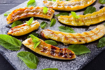 Halves of grilled bananas with powdered sugar on a black kitchen board and shale stone. Baked banana dessert with mint on black background table. Close up view