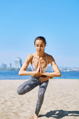 Young slim sports woman makes yoga balance exercises on the sand beach with city background against blue sky and sea. Young pretty fitness lady mixed race of Asian Caucasian ethnicity outdoors on the