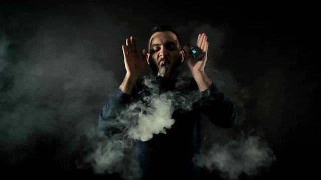 Bearded man vaping one big ring of steam with an electronic cigarette, moves forward to camera and repels circle with hand on black isolated background in slow motion in professional dark studio.