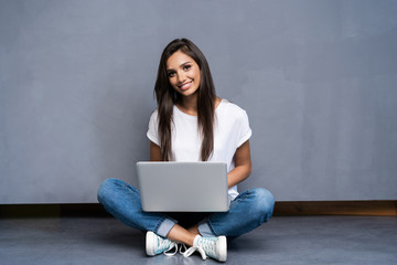 Happy young woman sitting on the floor with crossed legs and using laptop on gray background.