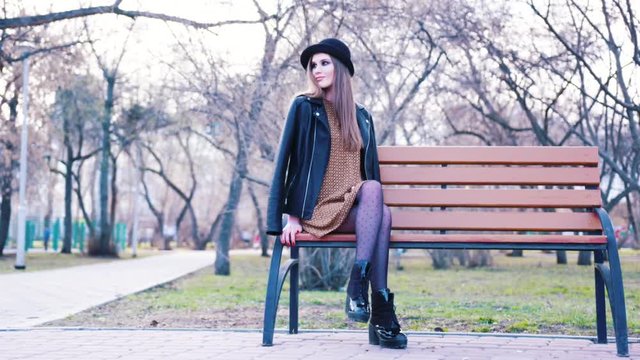 Attractive young woman in fashion dress, black hat, ankle boots and leather jacket sitting alone on the bench in park in warm spring day. Action. Urban modern style