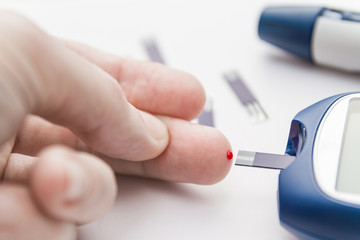 Man brings your finger to the test strip of the blood glucose meter during checking blood sugar level. Diabetes concept. Lancet pen and test strips on the background. Closeup, selective focus