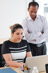 Obraz premium Asian young secretary sitting at the table and typing on laptop with serious male leader standing near by and watching her work