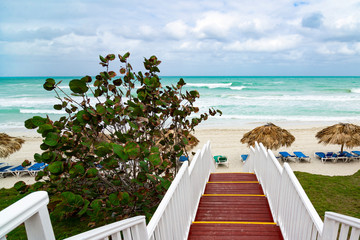 Naklejka premium Beautiful wooden ladder to the ocean beach. Beach with straw umbrellas and sun-loungers. Cloudy sky and blue water