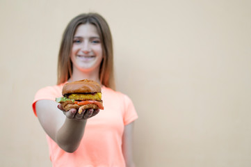 young teen girl stands against the orange wall in an orange T-shirt and holds a big burger in her hand, she brings the burger to the camera and smiles