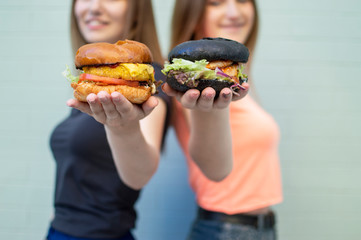 young beautiful girls-students stand near the blue wall, hold burgers and smile, they are happy and show cheeseburgers on camera