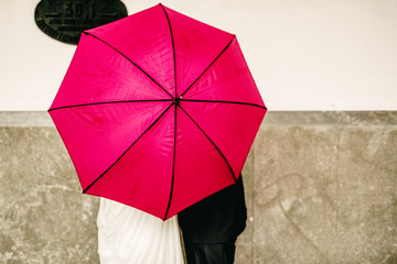 Couple in love hiding behind a big red umbrella while they kiss shyly hiding to keep their privacy and intimacy.