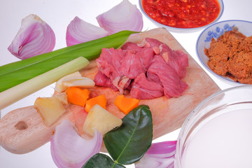 Food preparation detail - fresh red beaf meat on cutting board  with coconut milk , onion, garlic, lemon grass, ginger , turmeric, kerisik, chili and leaf turmeric