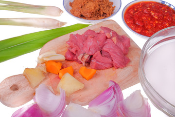 Food preparation detail - fresh red beaf meat on cutting board  with coconut milk , onion, garlic, lemon grass, ginger , turmeric, kerisik, chili and leaf turmeric