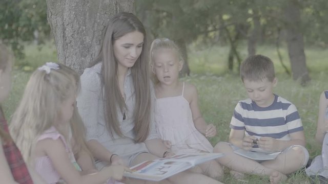 V-log. Young woman at work as educator reading book to boys and girls in park