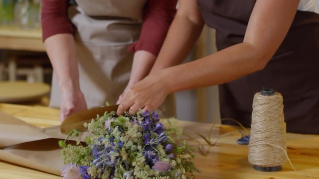 Tilting close-up shot of two happily smiling blonde florists working together in flower shop on wooden table and wrapping delicate fresh wildflower bouquet in plain brown paper, tying it with twine