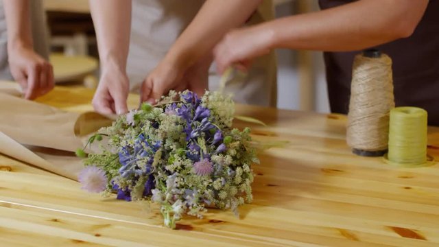 Close-up hands shot of three florists working together in brightly lit flower shop on wooden table, wrapping exquisite fresh wildflower bouquet in plain brown packaging paper and tying it with twine