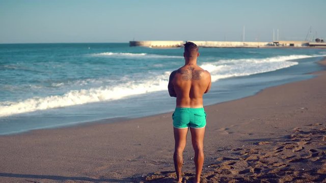 Tattooed bodybuilder sexy male coach at the beach. He stand and looks at ocean water