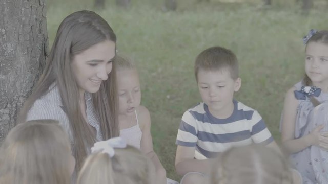 V-log. Young woman at work as educator reading book to boys and girls in park