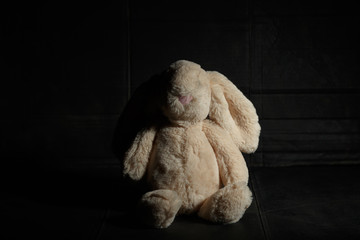 Soft toy bunny on a dirty dark background in the beam of hard light with shadow. Lost childhood...
