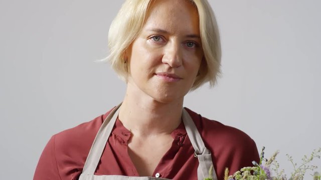 Tilting portrait shot of blonde middle-aged experienced florist, dressed in rusty red shirt and apron, posing against beige-grey wall with lovely bespoke wildflower bouquet and smiling modestly