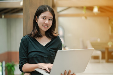 Closeup Asian beautiful woman Put on a black shirt and stand against the wooden pillar in the middle of the house. Use a laptop computer to work happily