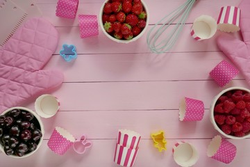 Summer berries cakes. Raspberries, strawberries, sweet cherry berries, muffin cups, whisk, cookie cutters on a soft pink wooden background.Berry baking concept.
