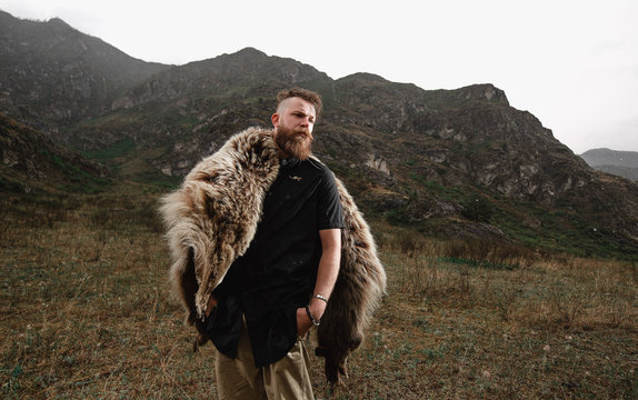 a serious and strict man with a beard and mustache in a black T-shirt and a bear skin stands at a cliff, a mountain.