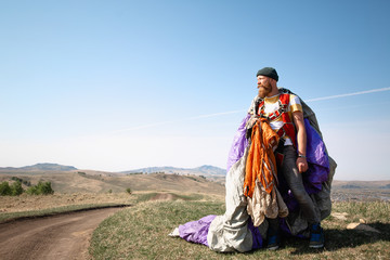 Fototapeta premium man with beard mustache striped white-yellow T-shirt and a green hat jeans with a red backpack. Carries a purple gray parachute on background of the mountains, a tattoo on his arm