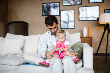 one year old blonde daughter sits on her father's lap on the couch at home