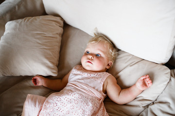 smiling one year old blond girl in a pinkish dress is lying on the sofa at home
