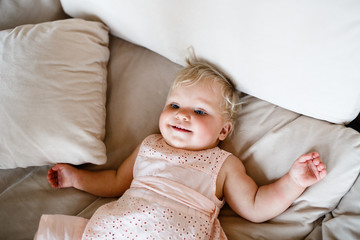 smiling one year old blond girl in a pinkish dress is lying on the sofa at home