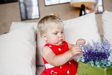 Indoor portrait of 1 year happy blond girl in a red jumpsuit playing with flower