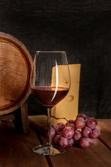 A glass of red wine with a wine barrel, a large piece of swiss cheese, and a bunch of grapes on a dark rustic background, low key photo with copyspace